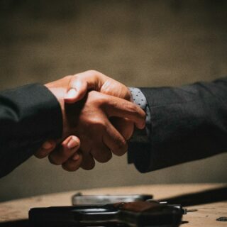 Two people in suits shake hands over a table, with a handgun resting in the foreground, suggesting a deal or confrontation.