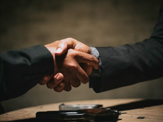 Two people in suits shake hands over a table, with a handgun resting in the foreground, suggesting a deal or confrontation.