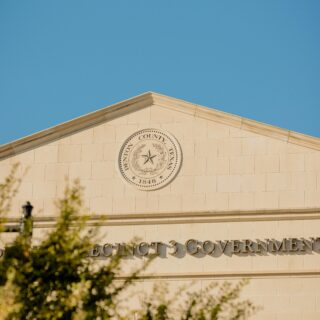 Beige stone government building with a circular seal and the word GOVERNMENT along the facade, blue sky behind and trees in the foreground.