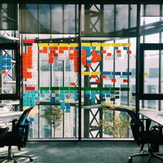 Open-plan office with a glass wall covered in colorful sticky notes in neat rows; two swivel chairs frame the scene.