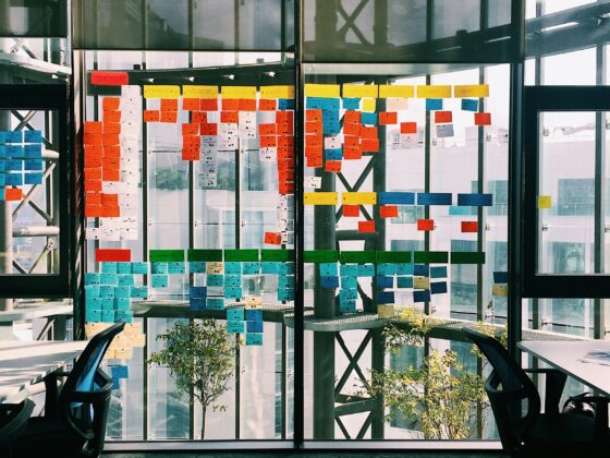 Open-plan office with a glass wall covered in colorful sticky notes in neat rows; two swivel chairs frame the scene.
