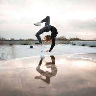 Person in a black top and gray jeans balancing on hands on a rooftop, legs bent in a gymnastic pose, with a cloudy sky behind and their reflection in a puddle