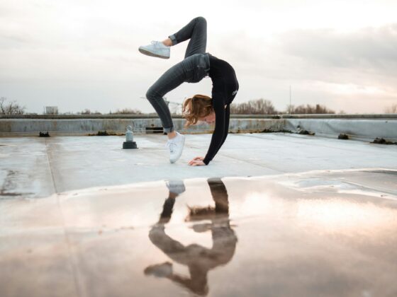 Person in a black top and gray jeans balancing on hands on a rooftop, legs bent in a gymnastic pose, with a cloudy sky behind and their reflection in a puddle