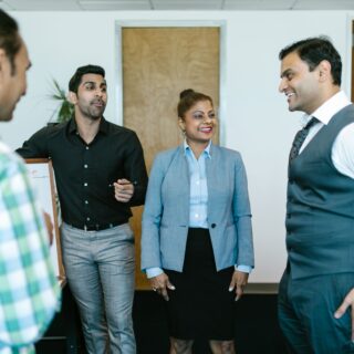 Group of four professionals in an office, engaged in conversation and smiling around a desk. One man in a black shirt gestures as others listen.