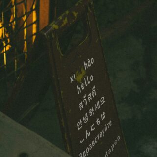 Tilted brown sign with white multilingual greeting text (e.g., 'hello') outdoors at night beside a lit fence and stone railing.