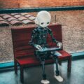 Humanoid robot with a white head sitting on a wooden bench, holding a magazine or booklet on its lap amid an outdoor setting.