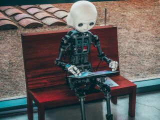 Humanoid robot with a white head sitting on a wooden bench, holding a magazine or booklet on its lap amid an outdoor setting.