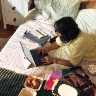 Person sits on a bed typing on a laptop, surrounded by notebooks, snacks, and a backpack in a cozy bedroom.