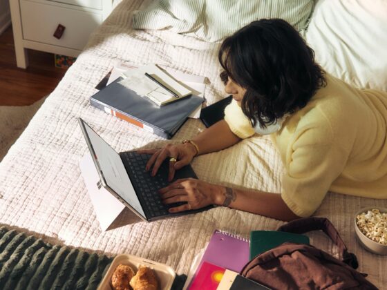 Person sits on a bed typing on a laptop, surrounded by notebooks, snacks, and a backpack in a cozy bedroom.