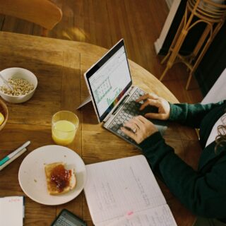 Person typing on a small laptop at a wooden breakfast table with a cereal bowl, toast, and juice nearby.