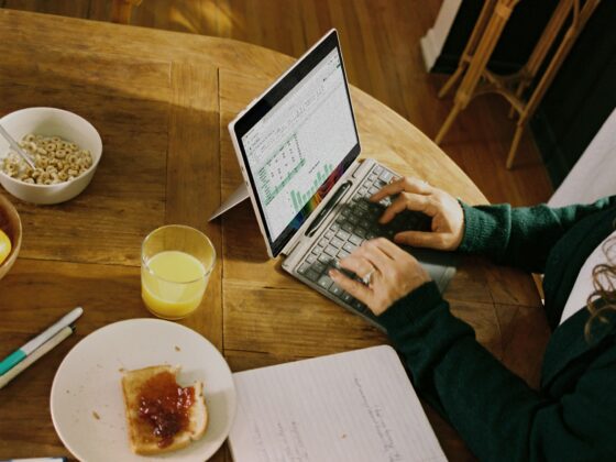 Person typing on a small laptop at a wooden breakfast table with a cereal bowl, toast, and juice nearby.