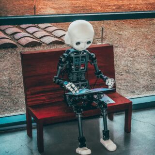 White-headed humanoid robot sits on a wooden bench outdoors, looking down at a magazine or booklet in its hands