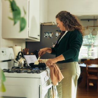 Woman standing at a gas stove, cooking, with a recipe tablet open on the counter and a kettle nearby in a bright kitchen.
