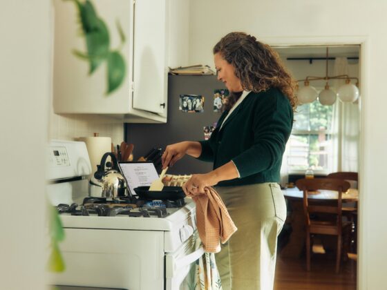 Woman standing at a gas stove, cooking, with a recipe tablet open on the counter and a kettle nearby in a bright kitchen.