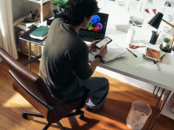 Person seated at a white desk, looking at a phone over a notebook with a laptop nearby and colorful screen. A workspace: person texting on a phone at a cluttered desk with papers, pens, and wall notes.