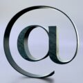 Large metallic @ symbol sculpture twisting in the air against a pale blue gradient background.