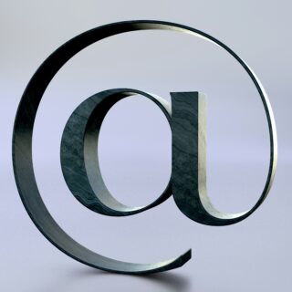 Large metallic @ symbol sculpture twisting in the air against a pale blue gradient background.