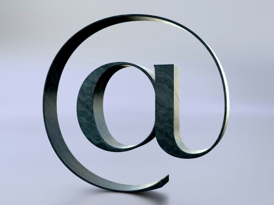 Large metallic @ symbol sculpture twisting in the air against a pale blue gradient background.