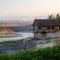 Thatched-roof cabin on stilts beside a river, with rocky banks and distant hills at dusk; calm, scenic landscape.