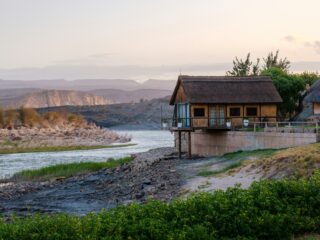 Thatched-roof cabin on stilts beside a river, with rocky banks and distant hills at dusk; calm, scenic landscape.