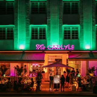 Night view of a restaurant facade with green uplighting and a pink neon sign reading 'The Carlyle', outdoor dining with people beneath.