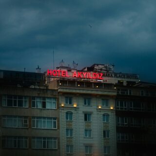 Red neon sign reading 'HOTEL AKYILDIZ' atop a multi-story building under a cloudy, evening sky at dusk.