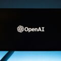OpenAI logo illuminated on a dark screen mounted outdoors against a blue sky backdrop