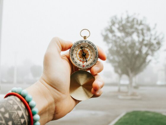 Close-up of a hand holding a compass outdoors, with beaded bracelets on the wrist visible in the foreground.