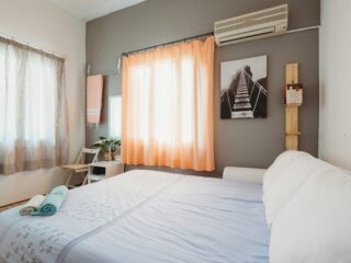 Bright bedroom with a bed in the foreground, light blue bedding with stars, and peach curtains by a large window.