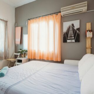 Bright bedroom with a bed in the foreground, light blue bedding with stars, and peach curtains by a large window.