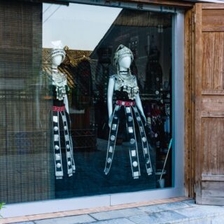 Mannequins wearing ornate silver jewelry and black embroidered outfits with headdresses in a storefront window, wooden doors visible on the right.