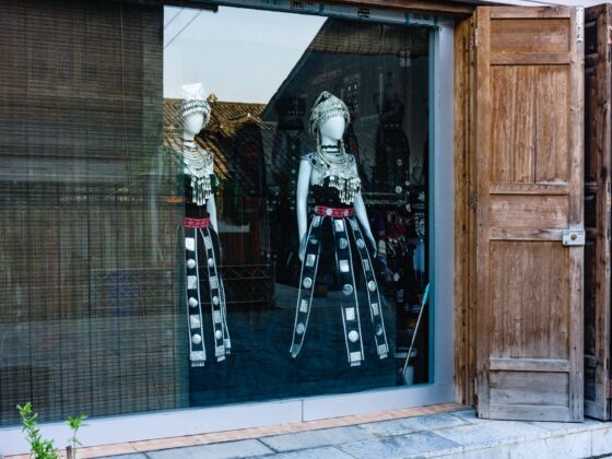 Mannequins wearing ornate silver jewelry and black embroidered outfits with headdresses in a storefront window, wooden doors visible on the right.