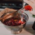 Hands pouring red liquid from a glass bottle into a metal mixing bowl on a kitchen counter, with a braided spoon resting across the bowl.