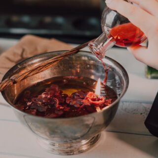 Hands pouring red liquid from a glass bottle into a metal mixing bowl on a kitchen counter, with a braided spoon resting across the bowl.