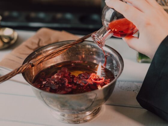 Hands pouring red liquid from a glass bottle into a metal mixing bowl on a kitchen counter, with a braided spoon resting across the bowl.
