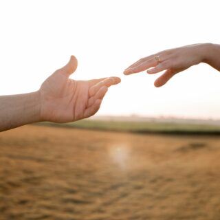 Two hands reaching toward each other over a sunlit beach, symbolizing connection or support, with one hand wearing a gold ring