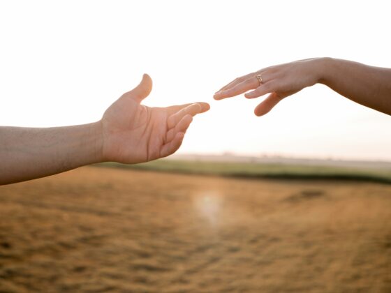 Two hands reaching toward each other over a sunlit beach, symbolizing connection or support, with one hand wearing a gold ring