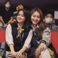 Two smiling girls in school uniforms sitting on red seats at an indoor event.
