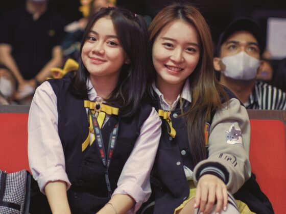 Two smiling girls in school uniforms sitting on red seats at an indoor event.
