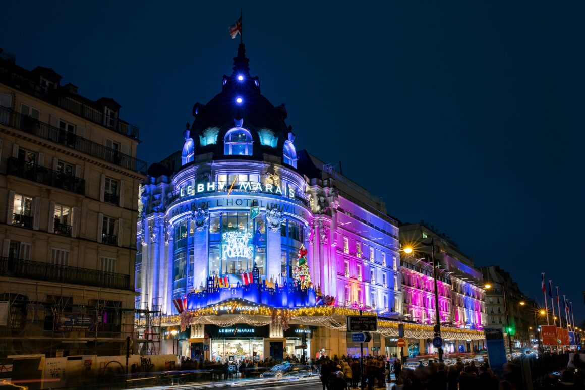 Nighttime facade of Le BHV Marais department store in Paris, illuminated in blue and pink; crowds at street level and festive lights above the entrance