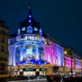 Nighttime facade of Le BHV Marais department store in Paris, illuminated in blue and pink; crowds at street level and festive lights above the entrance