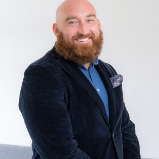 Portrait of a bald man with a red beard, wearing a navy blazer and blue shirt, smiling at the camera.