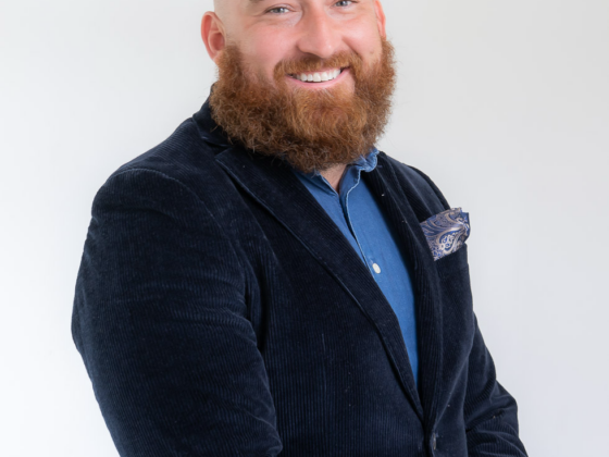 Portrait of a bald man with a red beard, wearing a navy blazer and blue shirt, smiling at the camera.
