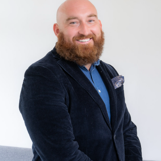Portrait of a bald man with a red beard, wearing a navy blazer and blue shirt, smiling at the camera.