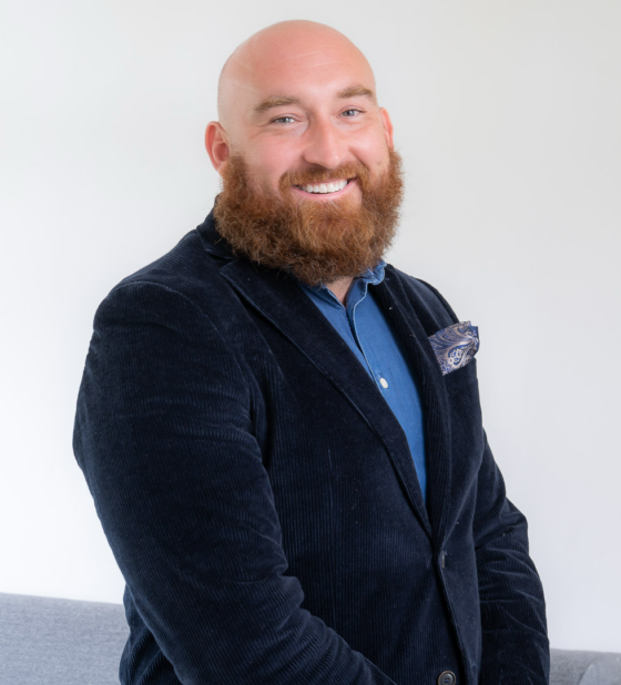 Portrait of a bald man with a red beard, wearing a navy blazer and blue shirt, smiling at the camera.