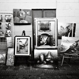 A street-side art display with paintings propped against a wall, including landscapes and buildings on wooden boards and a wire rack of smaller images left.