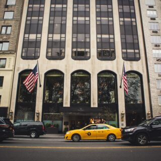 Urban city street with a tall beige building, large windows, and twoAmerican flags flanking the entrance; yellow taxi and parked cars in foreground.