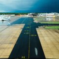 Wide airport runway and taxiways with planes at gates beneath a dark stormy blue sky and wet tarmac.