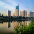 City skyline with tall blue glass skyscraper and other modern buildings along a river, reflecting in calm water; foreground greenery and trees frame the scene.