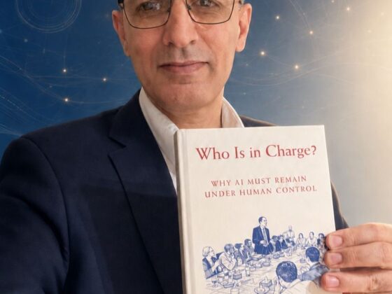 Man in a suit and glasses holding a white book titled 'Who Is in Charge?' about AI and human control.
